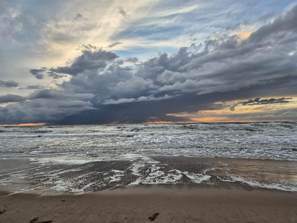 A wide storm cloud spread across the sky above a beach, with foam-streaked waves rolling onto flat wet sand.